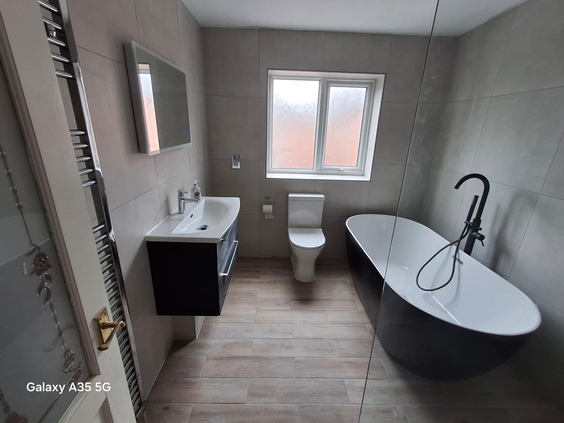 Modern fitted bathroom with large grey tiles, wall-hung black vanity, freestanding black bath with matt black tap and chrome heated towel rail